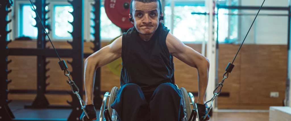 Foto von Anas. Er sitzt im Rollstuhl und zieht in einem Fitnessstudio an zwei Seilzügen. Er schaut angestrengt.