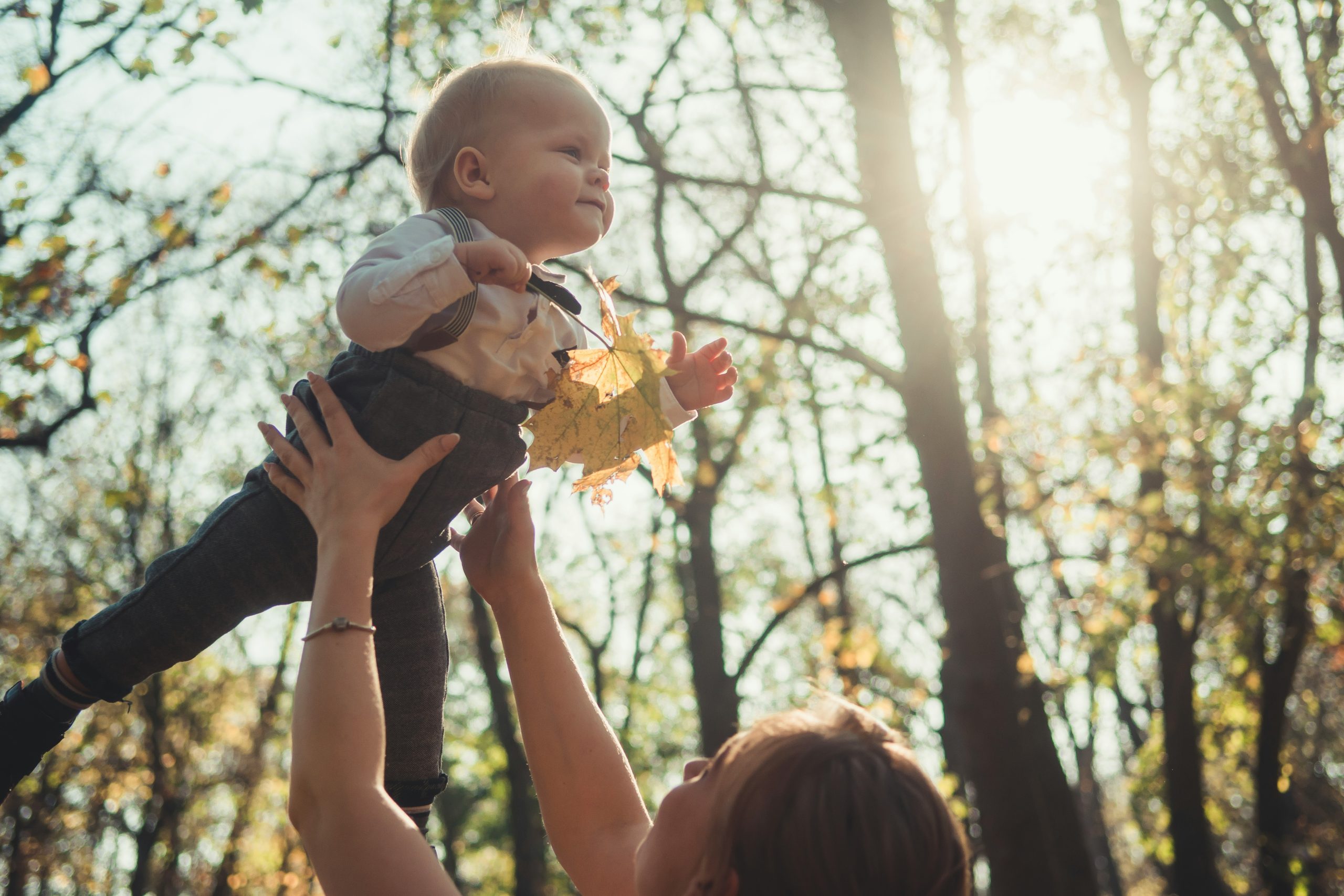 Weiß positionierte Person hält Baby hoch in die Luft, das Kind lacht, im Hintergrund ein Wald.