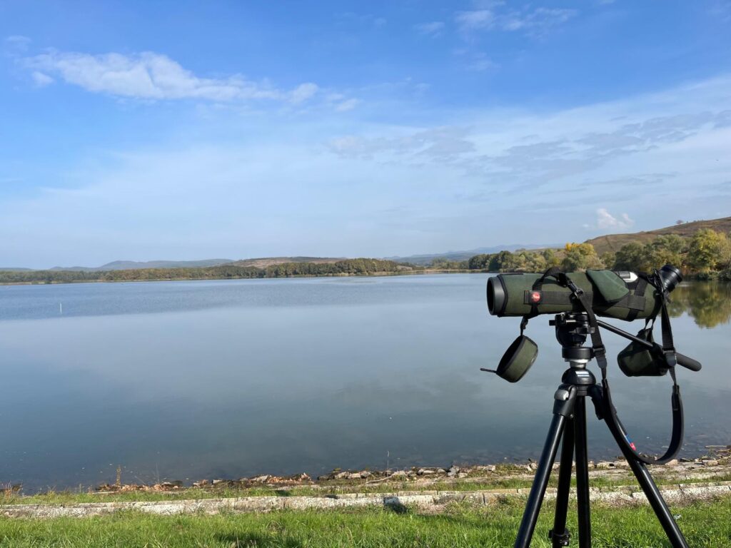 Eine professionelle Kamera steht auf einem Stativ im rechten Bildvordergrund auf einem Rasenstück, dahinter liegt ein See, im Hintergrund sind Bäume und Hügel zu sehen, der Himmel ist blau mit wenigen Wolken.
