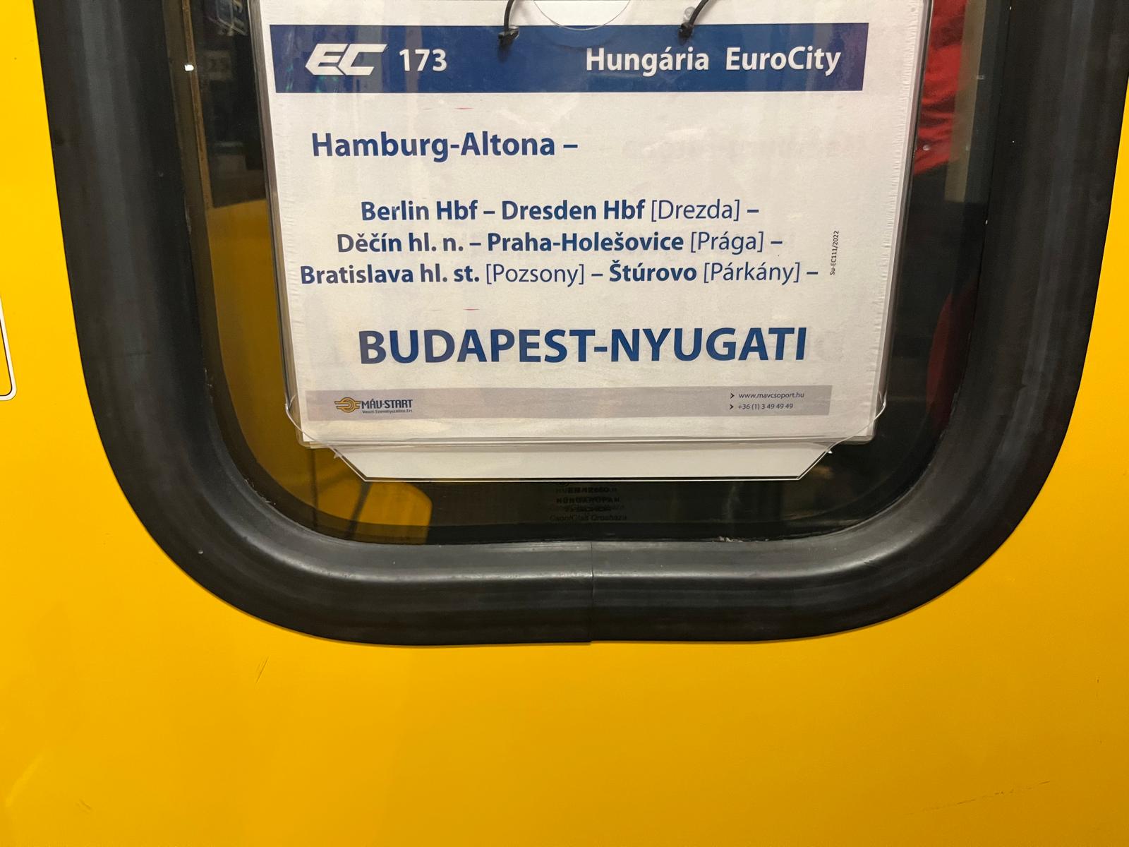An einem gelb umrahmten Zugfenster ist ein Schild mit der Aufschrift Hungaária EuroCity: Hamburg Altona - Budapest-Nyugati zu sehen.