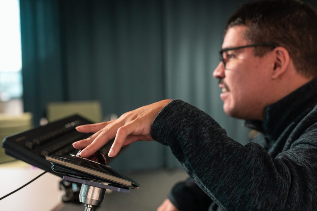 Foto von Sven Papenbrock. Er hat schwarze kurze Haare und trägt eine Brille. Er sitzt vor einem Bildschirm und arbeitet an einer erhöhten Tastatur.