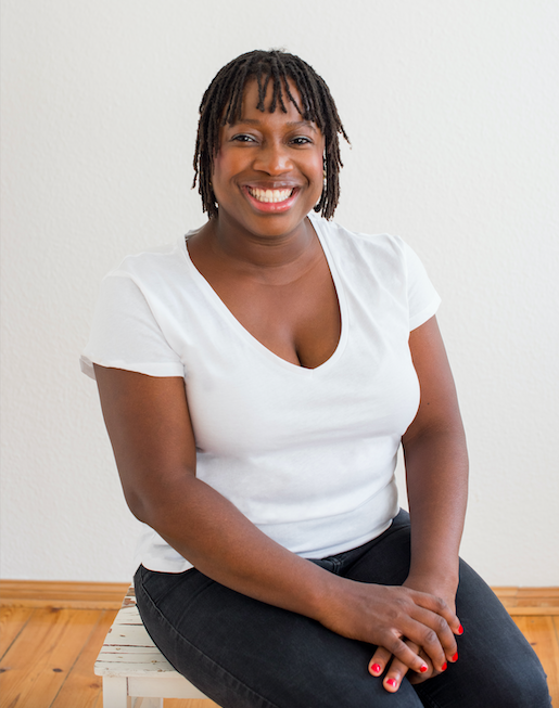 Die Autorin Olaolu Fajembola sitzt auf einem Holzhocker. Sie trägt ein weißes T-Shirt und eine schwarze Hose. Ihre Hände mit rot lackiertem Nagellack hat sie übereinandergelegt und schaut lächelnd in die Kamera.