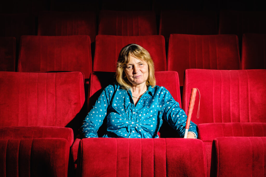 Eine Frau mit einer blauen Bluse und weißen Punkten sitzt auf einem roten Kinosessel. Der Ansatz ihres Langstockes ist zu sehen.