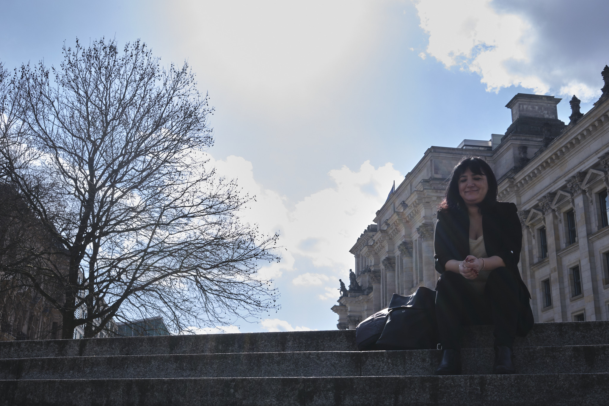 Gökay Akbulut sitzt im schwarzen Blazer, schwarzer Hose und hellem Shirt auf einer Treppe. Ihre Hände liegen auf den Knien ineinander, sie lächelt und hat die Augen geschlossen. Links im Bildhintergrund ist ein kahler Baum zu sehen, rechts der Bundestag.