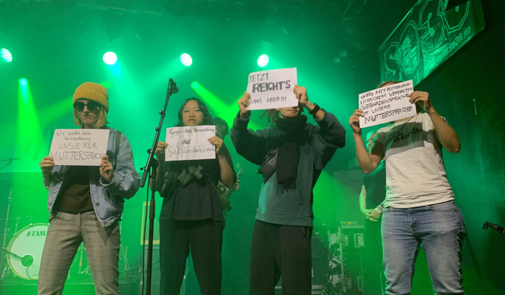 Vier Aktivist*innen von Deaf Performance Not stehen auf einer grün erleuchteten Bühne und halten Plakate hoch, wo beispielsweise darauf steht: „Gegen die Vermarktung unserer Muttersprache“.