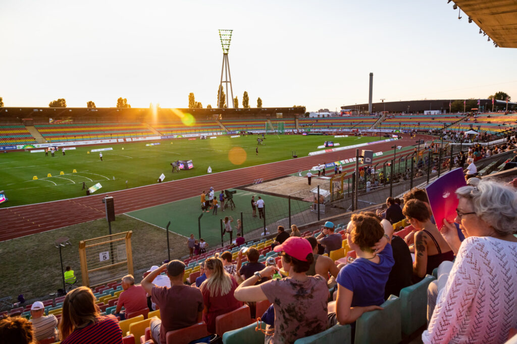 Blick in den Ludwig-Jahn-Sportpark. Auf den Tribünen sitzen Menschen, die einer Leichtathletik-Veranstaltung zuschauen. Im Hintergrund geht die Sonne unter.