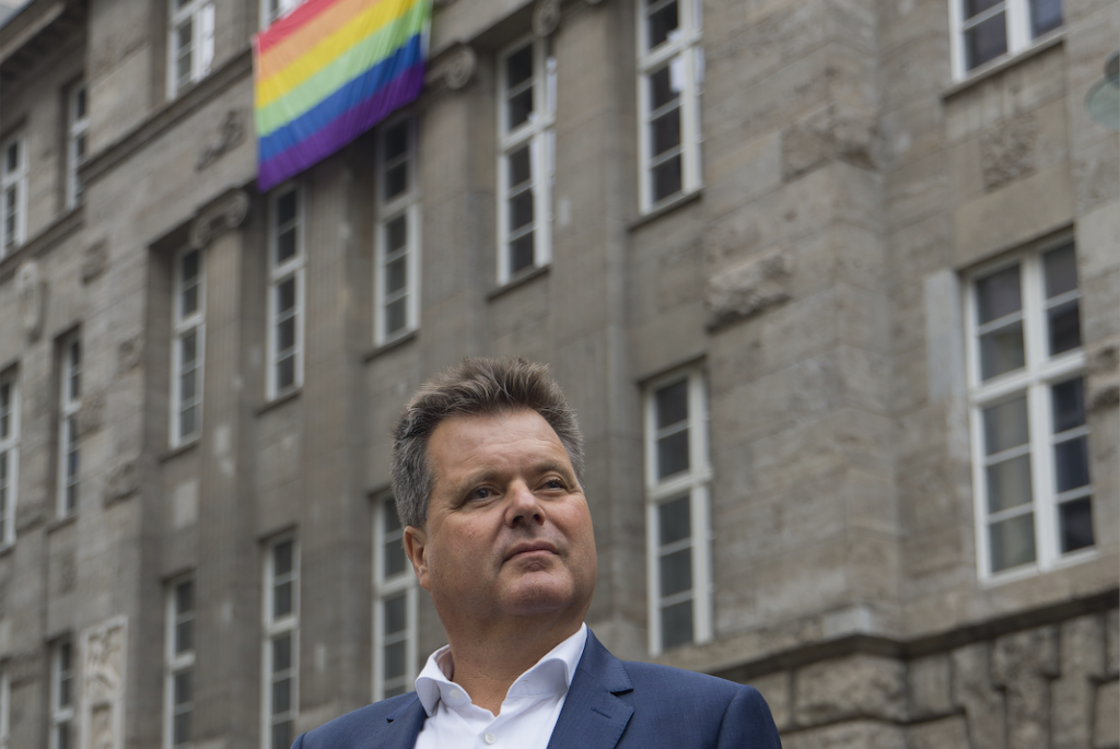 Jürgen Dusel trägt ein weißes Hemd und einen blauen Blazer. Hinter ihm ist ein altes Steingebäude zu sehen - aus dem einen Fenster hängt eine Regenbogenflagge.