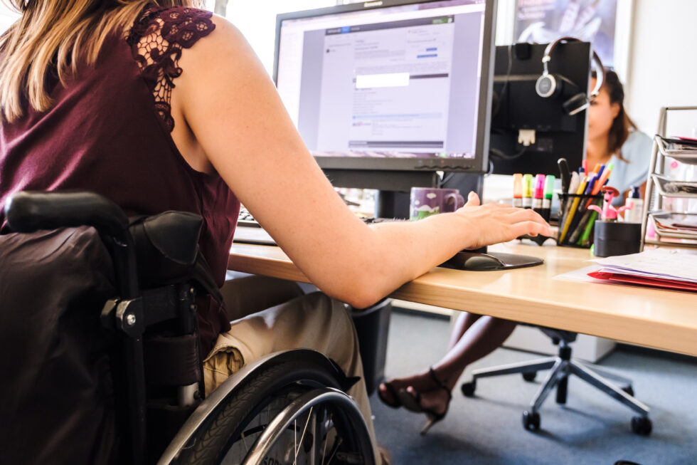 Eine Frau im Rollstuhl sitzt an einem Schreibtisch und arbeitet an einem Computer.