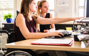 Eine Frau im Rollstuhl zeigt einer anderen Frau ohne Behinderung etwas auf einem Computerbildschirm.