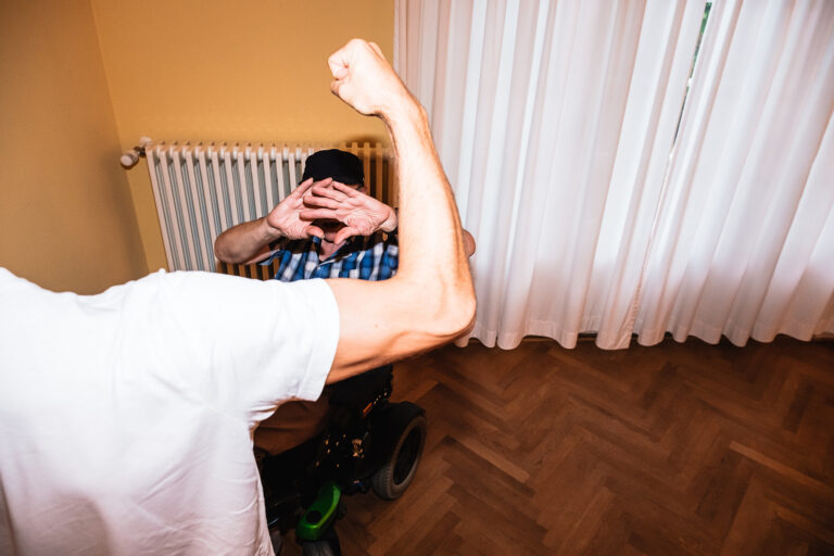 Eine erhobene Faust vor einer Person die im Rollstuhl sitzt und schützend die Hände vor das Gesicht hält.
