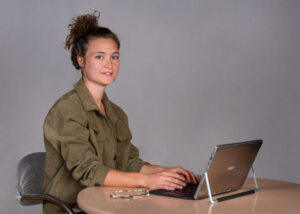 Eine Frau sitzt vor einer grauen Wan an einem Tisch vor einem Laptop. Sie hat schwarze, zum Dutt gebundene Haare und schaut in die Kamera.