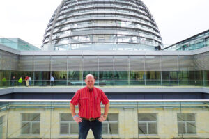Ein Mann im roten Hemd steht vor der Kuppel des Reichstages. Er hat die Hände in die Hüften gestemmt und blickt in die Kamera.