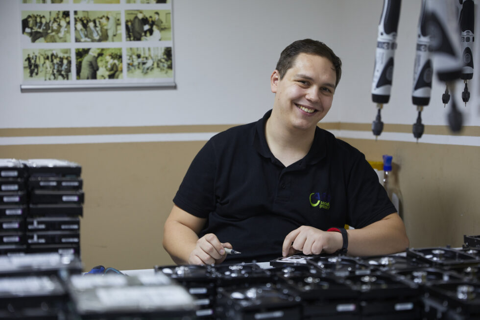 Ein Mann mit Behinderung trägt ein schwarzes T-Shirt, steht in einer Technik-Werkstatt und lächelt in die Kamera.