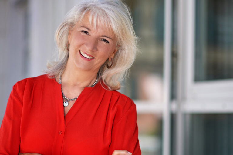 Eine Frau mit blonden langen Haaren und roter Jacke lächelt in die Kamera.