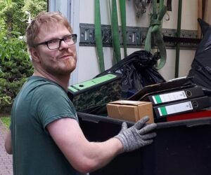 Ein Mann mit behinderung steht vor einem Müllcontainer, in den er Sachen wirft. Er lächelt in die Kamera.