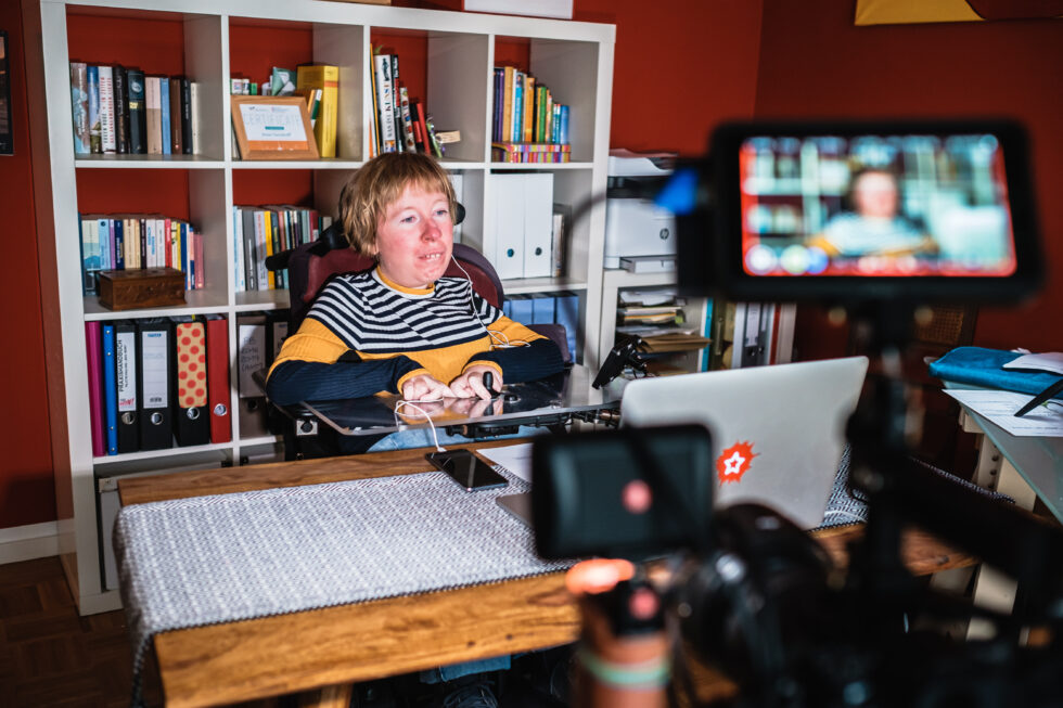 Anne Gersdorff sitzt im Elektrorollstuhl hinter ihrem Schreibtisch und wird von Kameras gefilmt.