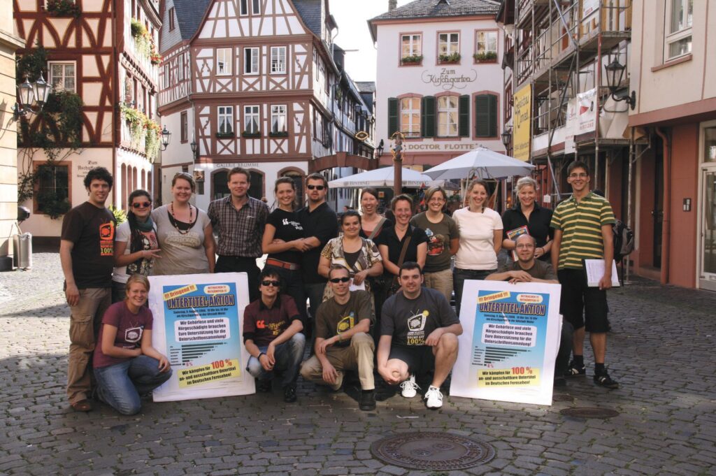 Eine Gruppe von 18 Menschen steht und kniet in einer Fußgänger*innenzone mit Fachwerkhäusern im Hintergrund.