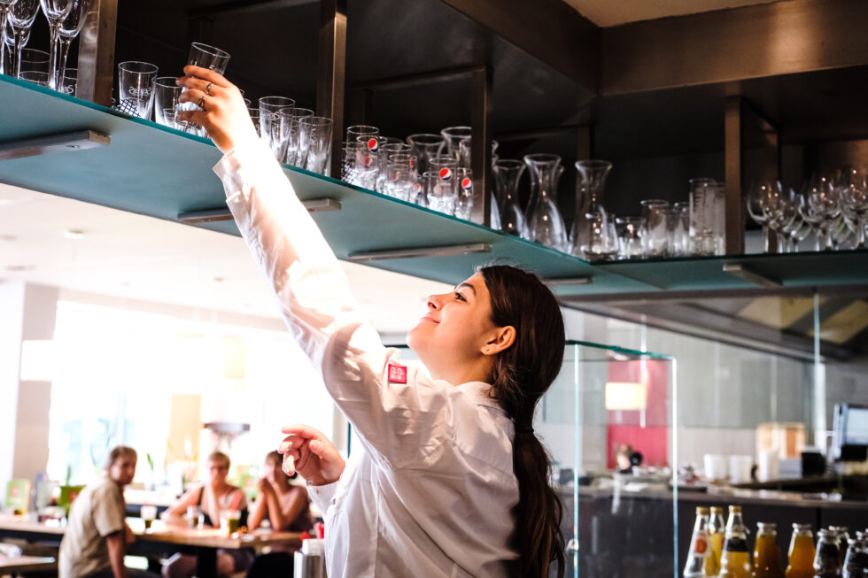 Eine Frau mit Behinderung greift nach Gläsern, die an einer Bar an der Decke hängen.