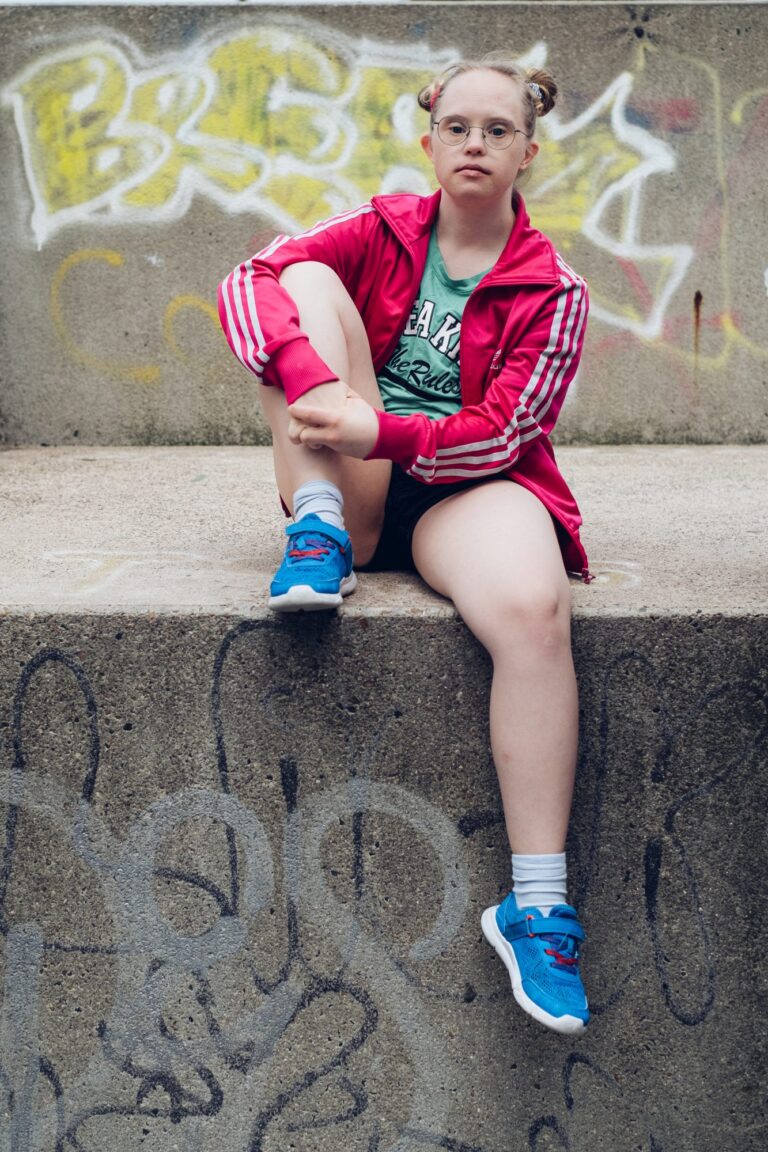 Neele Buchholz sitzt auf einer Betonmauer. Sie trägt eine kurze, schwarze Hose, ein grünes Shirt, eine pinke Trainingsjacke und blaue Turnschuhe.