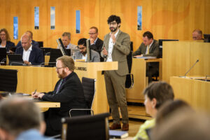 Bijan Kaffenberger steht im hessischen Landtag am Pult und hält eine Rede. Er hat schwarze Haare, einen Bart und trägt eine schwarze Brille und einen dunkel beigen Anzug mit weißem Hemd.