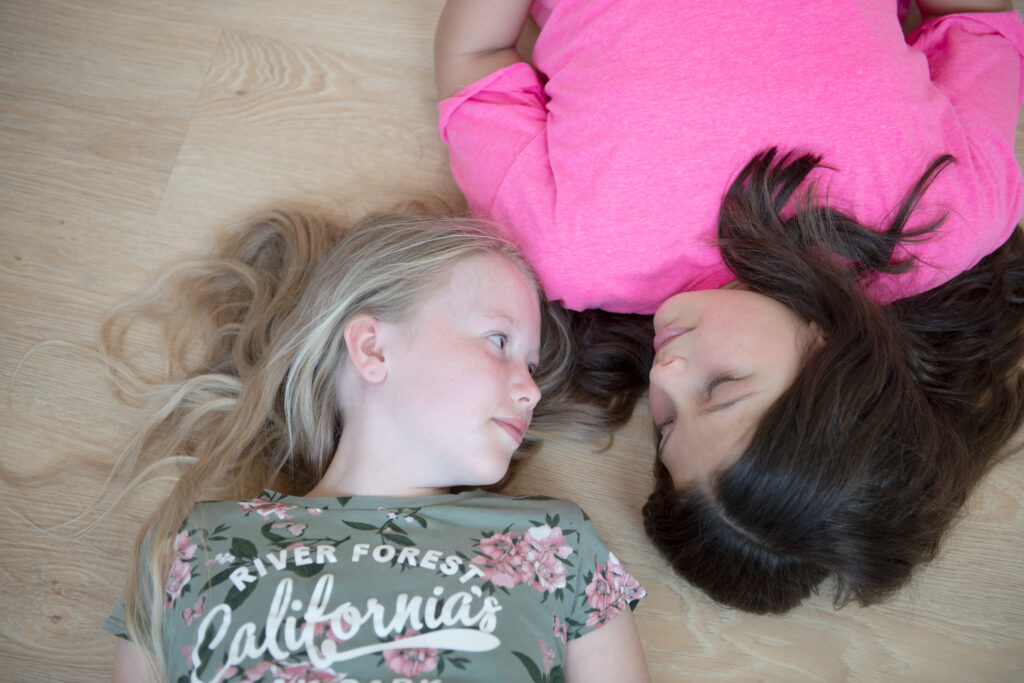 Zwei Grundschulkinder aus der Vogelperspektive. Sie liegen nebeneinander, die Füße in entgegengesetzte Richtungen und sehen sich an. Ein Mädchen hat längere blonde Haar und ein Shirt mit Schriftzug und Blumenmotiv, das andere hat dunkelbraune, längere Haare und trägt ein pinkes Shirt.