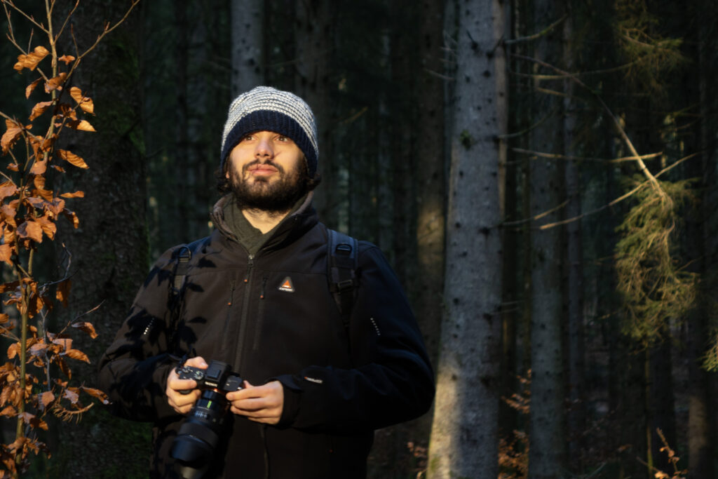 Manuel Stark steht, mit einer Spiegelreflexkamera in der Hand, angeleuchtet in einem dunklen Wald.