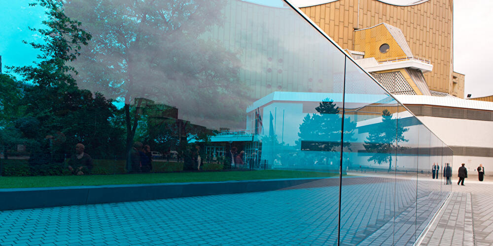 Im Vordergrund ist eine lange, transparente, blaue Glaswand. Im Hintergrund ist die Philharmonie zu sehen.