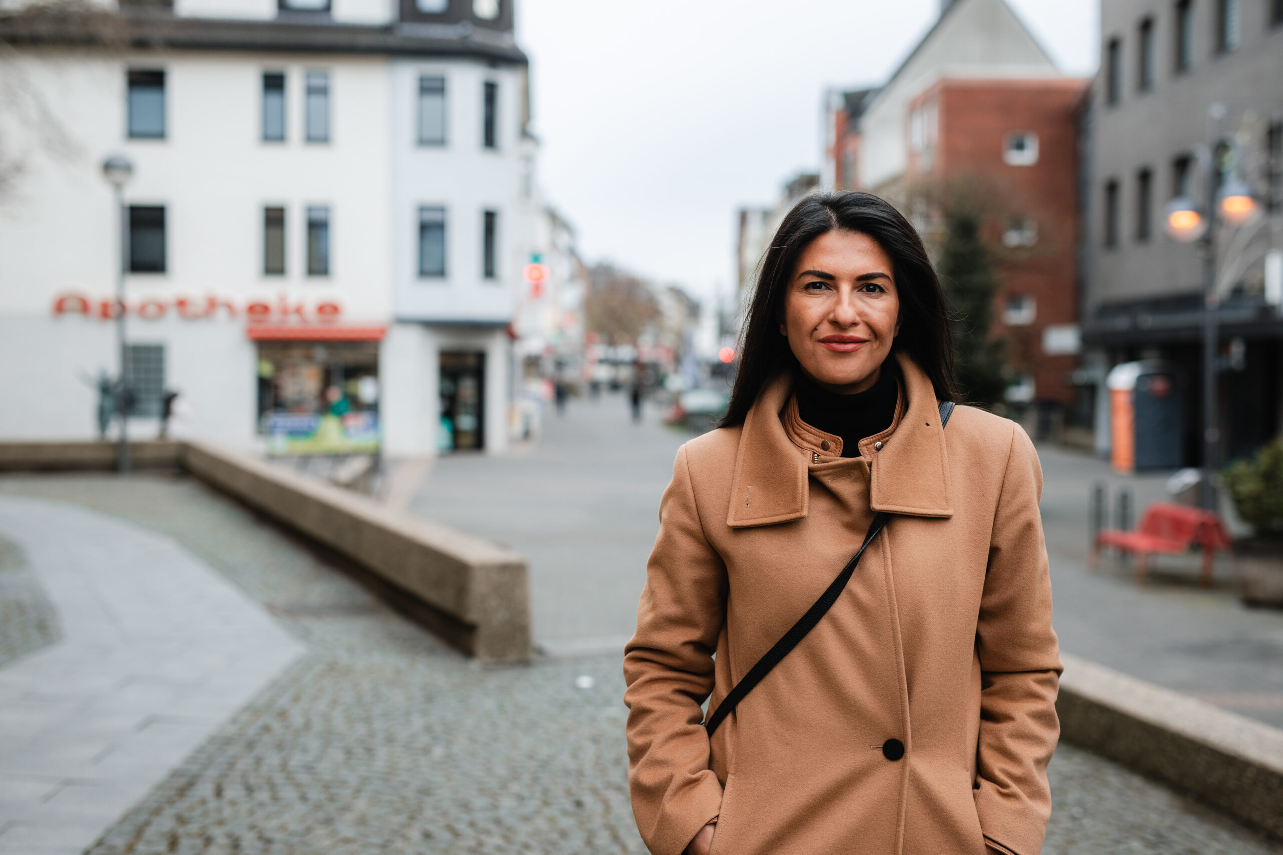 Serap Güler trägt einen hellbraunen Trenchcoat und eine schwarze Umhängetasche. Im HIntergrund ist eine Kleinstadt mit Apotheke und Marktplatz zu erkennen. Güler trägt ihre mittellangen, dunkelbraunen Haare offen.