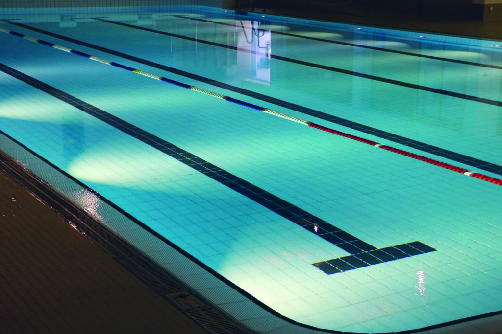 Ein leeres Schwimmbecken in einem Hallenbad, im Wasser spiegelt sich der Durchgang zu den Duschen.