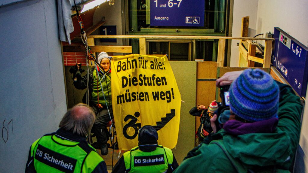 Eine Aktivistin im Rollstuhl hängt an einem Seil über einem Treppenzugang zu einem Bahngleis. Auf einem gelben Banner neben ihr steht „Bahn für alle. Die Stufen müssen weg.“ Darunter ist ein Rollstuhl-Symbol und ein Treppensymbol zu sehen. Neben ihr steht eine weitere Aktivistin. Auf den Stufen stehen zwei Personen vom Sicherheitspersonal der Bahn in Warnwesten. Daneben ist ein Fotograf zu sehen, der die Szene fotografiert.