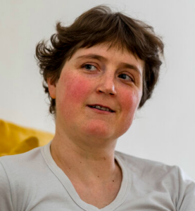 Portraitfoto einer weiß positionierten Frau mit kurzen, Braunen Haaren und weißem T-Shirt. Sie hat rote Backen, lächelt leicht und sieht nicht direkt in die Kamera.