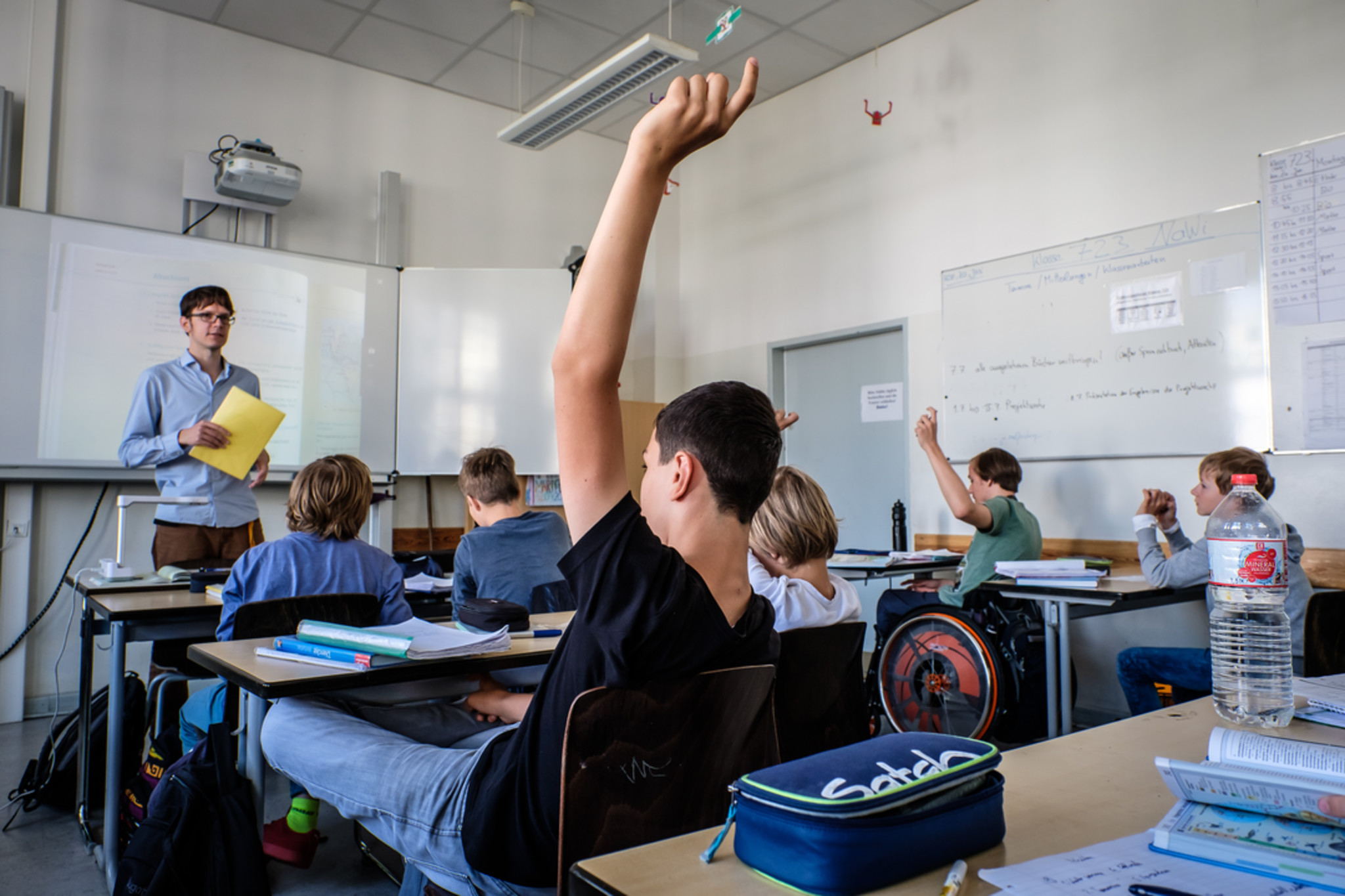 Blick von hinten in eine Schulklasse. Einige melden sich und eine Person sitzt im Rollstuhl.