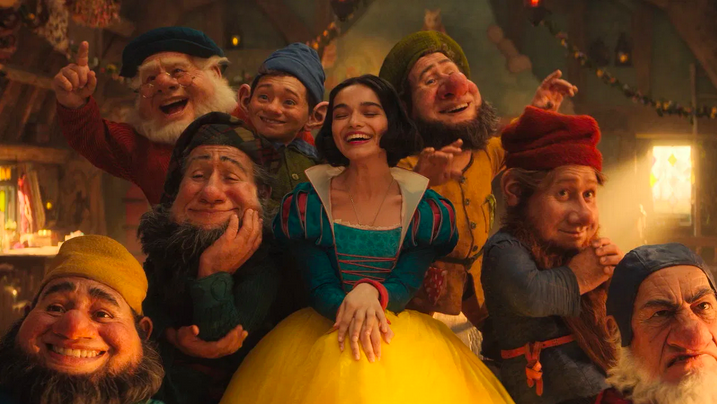 Screenshot aus der Neuverfilmung von Schneewittchen (2025). In der Mitte steht Rachel Zegler als Schneewittchen in einem türkis-gelben Ballkleid, neben ihr die animierten Zwerge. Sie sind weiß positioniert, tragen alle Bärte und haben große Nasen.