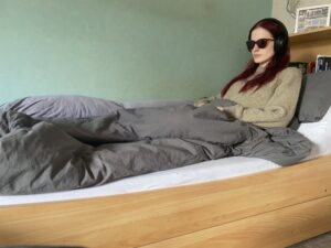 Eine Frau mit langen dunklen Haaren sitzt mit Sonnenbrille im Bett.