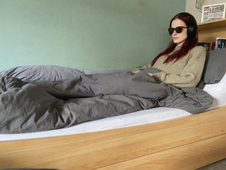 Eine Frau mit langen dunklen Haaren sitzt mit Sonnenbrille im Bett.
