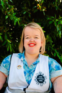 Porträt einer weiß positionierten, weiblich gelesenen Person, die lachend in die Kamera schaut. Sie hat schulterlanges, blondes Haar und trägt ein hellblau/weißes Hemd mit einer weißen Weste und roten Lippenstift. Im Hintergrund ist ein grüner Busch.