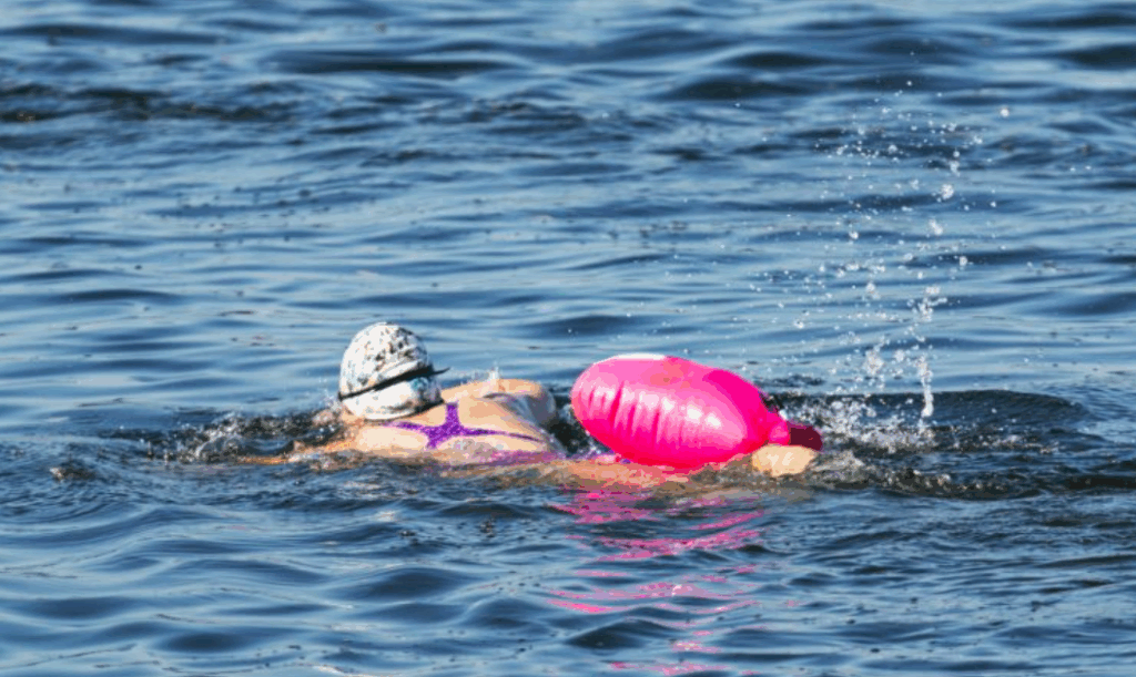 Weibliche, weiß gelesene Person mit Schwimmhaube und Schwimmbrille von hinten mit pinker Schwimmboje im See.