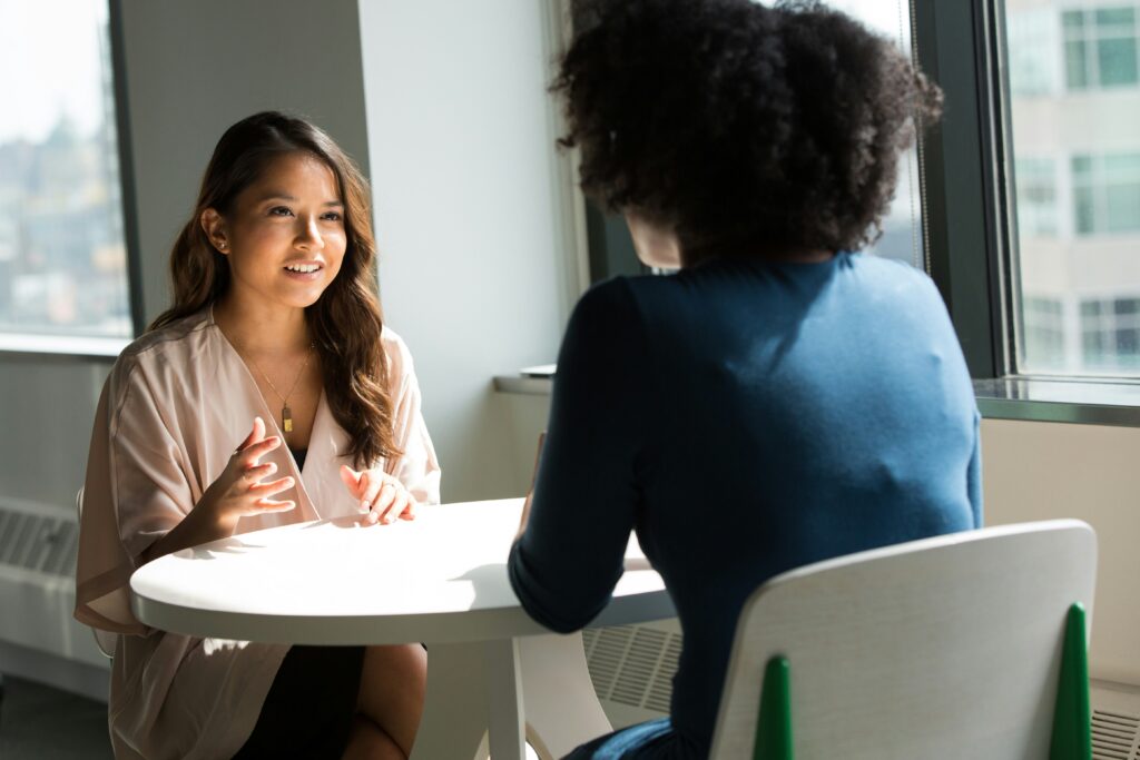 Zwei weiblich gelesene Personen sitzen an einem runden Tisch, die zur Betrachterin gewandte ist asiatisch gelesen, die vordere Person sitzt mit dem Rücken zur betrachtenden Person und trägt Afro-Haare. Beide unterhalten sich angeregt.