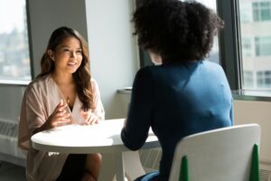 Zwei weiblich gelesene Personen sitzen an einem runden Tisch, die zur Betrachterin gewandte ist asiatisch gelesen, die vordere Person sitzt mit dem Rücken zur betrachtenden Person und trägt Afro-Haare. Beide unterhalten sich angeregt.