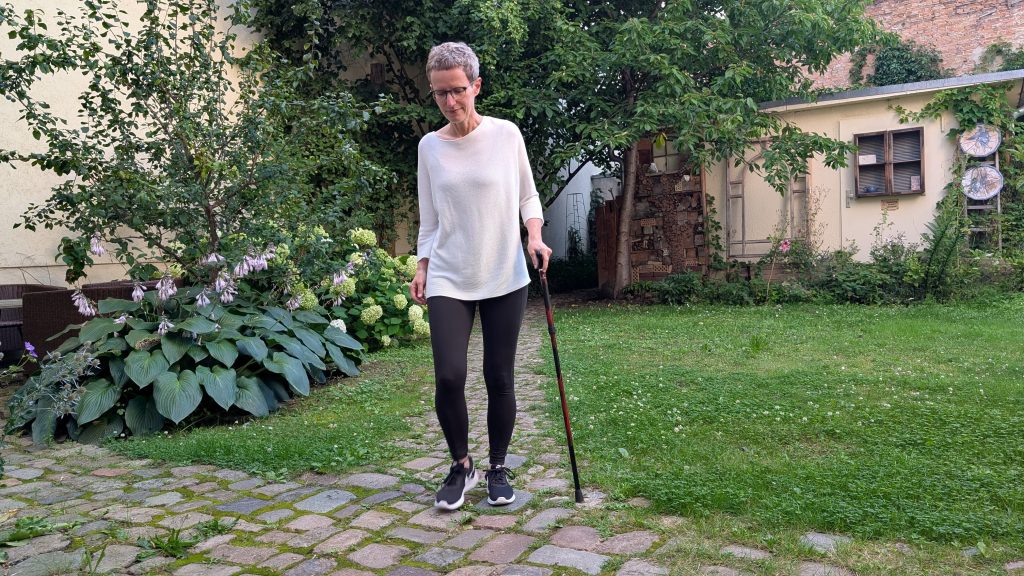 Eine Frau mit kurzen grauen Haaren läuft mit einem Gehstock über einen Weg im Garten.