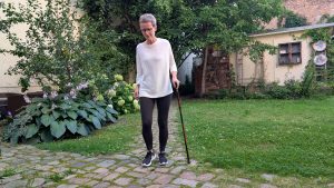 Eine Frau mit kurzen grauen Haaren läuft mit einem Gehstock über einen Weg im Garten.