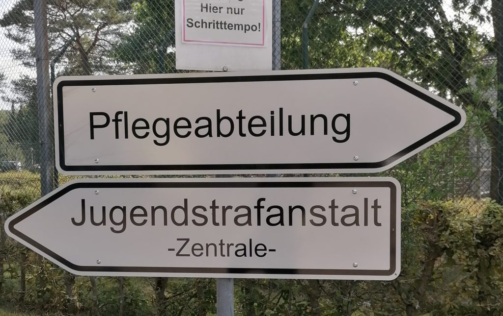 Zwei Verkehrsschilder. In der einen Richtung geht es zur Pflegeabteilung. In der anderen Richtung geht es zur Zentrale der Jugendstrafanstalt.