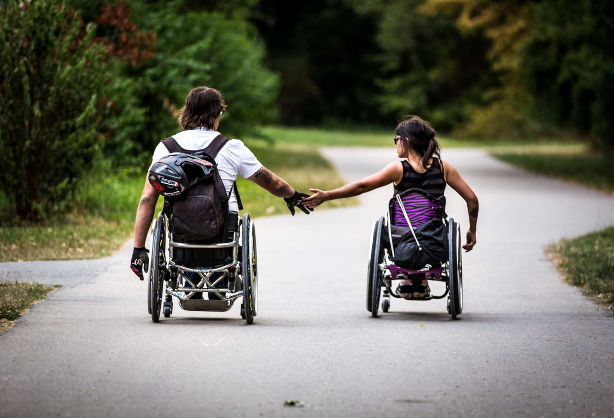 Zwei Menschen, die einen Rollstuhl nutzen, fahren auf einem geschlungenen Weg, rechts und links ist Parkfläche. Sie halten sich aus etwas Entfernung an den Händen.