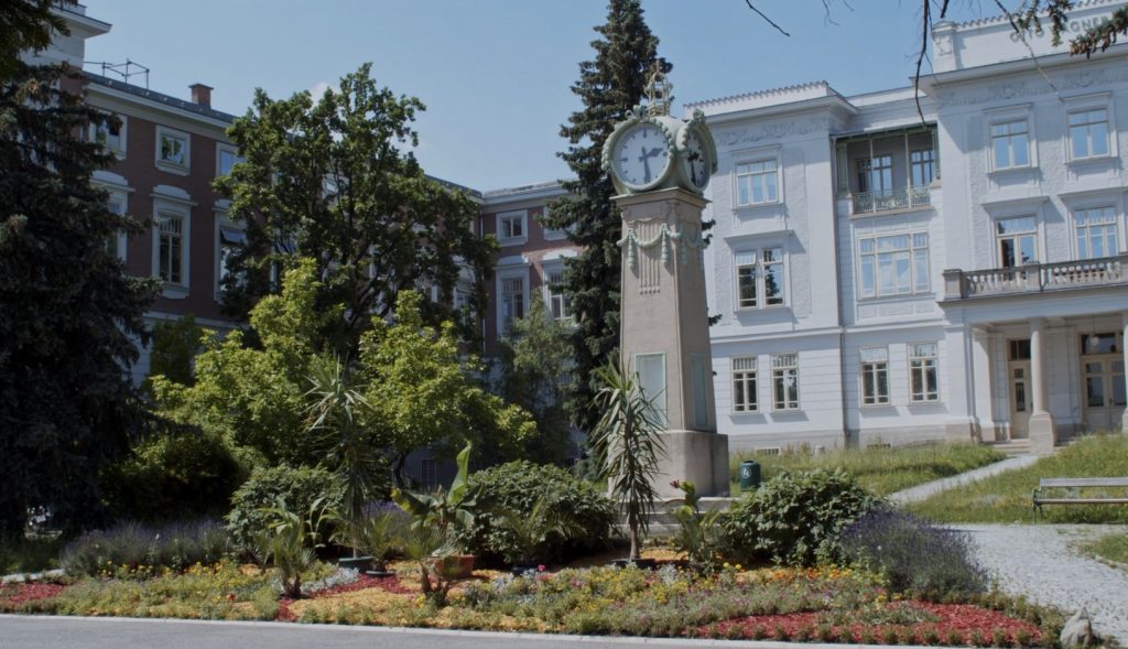 Im Hintergrund ist ein stattliches Klinikgebäude zu sehen, davor ein kleiner Park mit einer großen Standuhr.