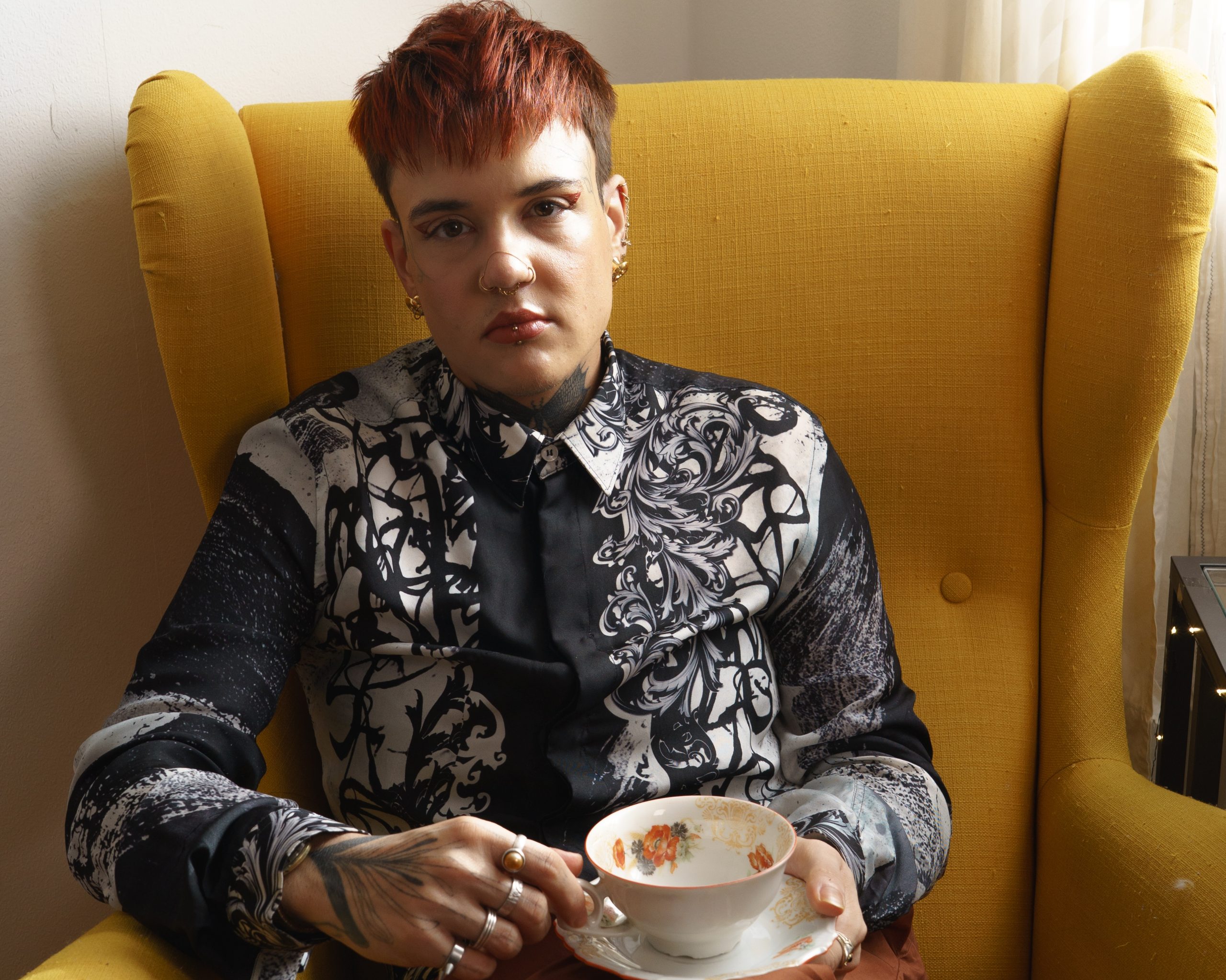 Eine Person mit kurzen Haaren sitzt in einem orangenen Sessel und hält eine Kaffeetasse in der Hand. Sie trägt ein schwarz-weißes Hemd mit Ornamenten und hält eine Teetasse in beiden Händen. Sie blickt in die Kamera.