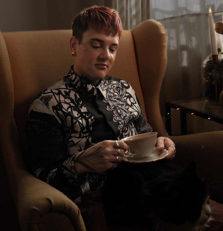 Eine Person mit kurzen Haaren sitzt in einem orangenen Sessel und hält eine Kaffeetasse in der Hand. Sie trägt ein schwarz-weißes Hemd mit Ornamenten und schaut auf die Teetasse, die sie in beiden Händen hält.