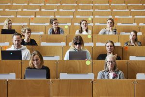 In einem Hörsaal sitzen 11 Personen. Eine weiblich gelesene Person erzählt etwas und die anderen Personen hören zu oder arbeiten an ihrem Laptop. Neben der Person, die spricht, sitzt eine weiblich gelesene Person, die eine schwarze Sonnenbrille trägt und auf ihren Laptop schaut. Das Bild ist sehr frontal aufgenommen und es wirkt fast die ein Gemälde durch die ganzen braunen uni-typischen Sitzreihen.