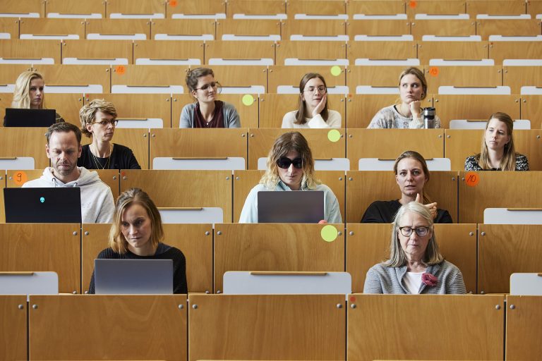 In einem Hörsaal sitzen 11 Personen. Eine weiblich gelesene Person erzählt etwas und die anderen Personen hören zu oder arbeiten an ihrem Laptop. Neben der Person, die spricht, sitzt eine weiblich gelesene Person, die eine schwarze Sonnenbrille trägt und auf ihren Laptop schaut. Das Bild ist sehr frontal aufgenommen und es wirkt fast die ein Gemälde durch die ganzen braunen uni-typischen Sitzreihen.