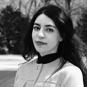 Schwarz-weiß Porträt einer Person mit langen, schwarzen Haaren, schwarzem Hemd mit hellem Jacket darüber. Sie trägt Lippenstift und lächelt leicht in die Kamera.