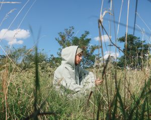 Das Foto wurde auf Augenhöhe aufgenommen und zeigt eine junge Frau mit Kopftuch, die in einem Feld sitzt. Sie trägt einen Kapuzenpullover, unter dem das Kopftuch sichtbar ist. Die Frau sitzt mit angewinkelten Beinen auf dem Boden, hält ein Handy in der Hand und richtet ihren Blick darauf. Umgeben ist sie von grünen Gräsern und Blumen, von denen einige im Vordergrund des Bildes unscharf zu sehen sind. Die obere Bildhälfte zeigt einen blauen Himmel mit einzelnen Wolken.